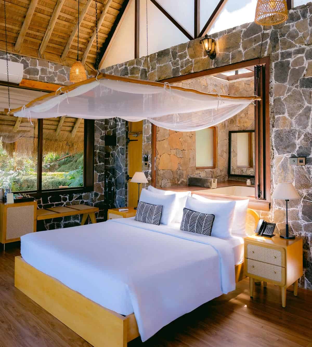 Rustic-chic hotel guestroom with stone walls, wood floors, a king bed and thatched ceiling