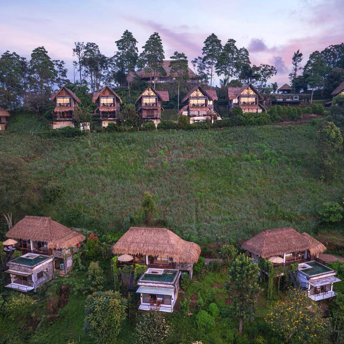 Thatch-roof chalets (some with private pools) are scattered across a green hillside, against a backdrop of a purple sky.
