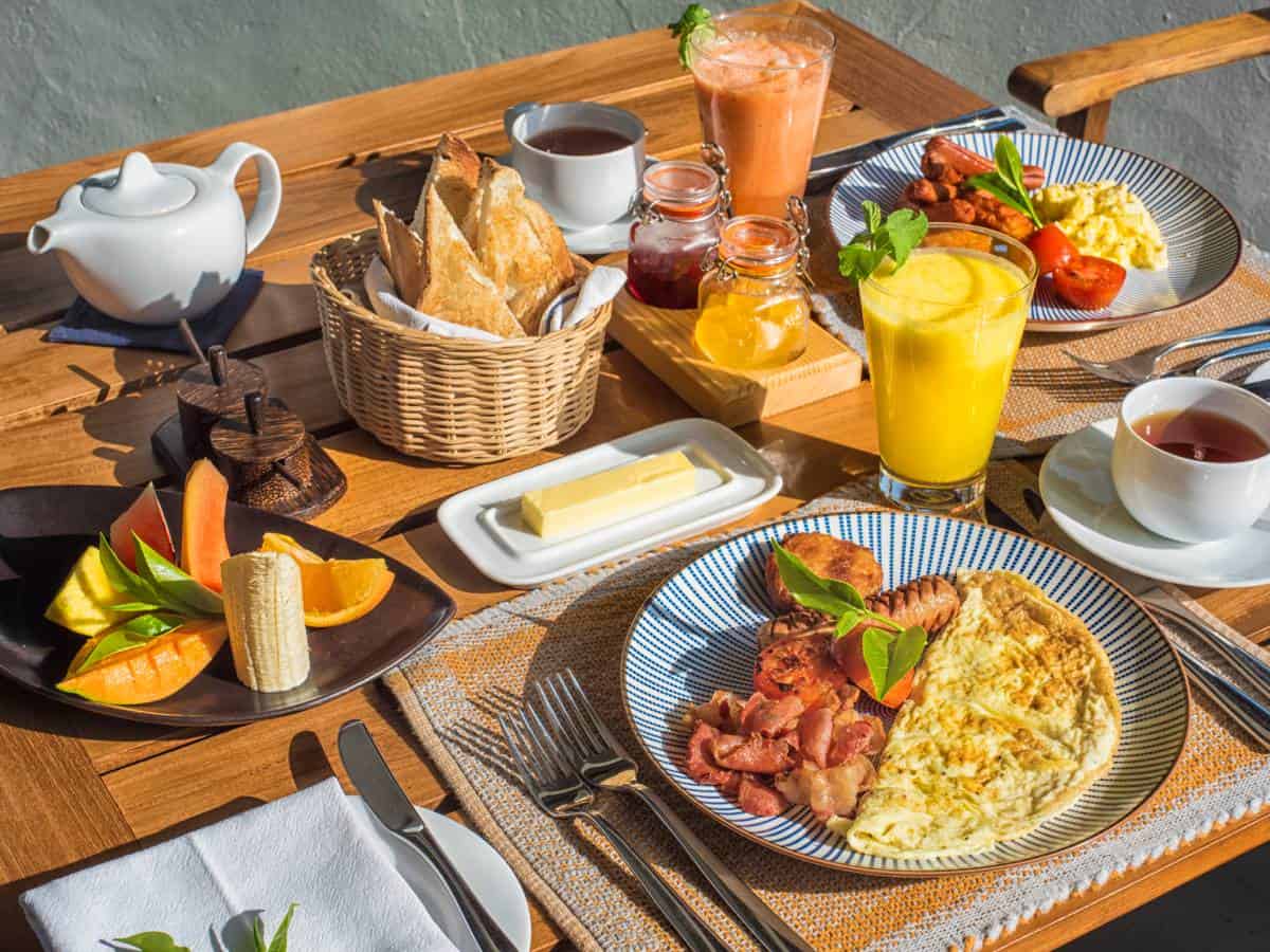 Breakfast table set with plates of omelettes, bacon and sausages; bowl of fruit and pot of tea